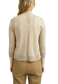 Femme portant un pull beige à manches longues et un pantalon marron, vue de dos avec les cheveux lâchés sur les épaules.