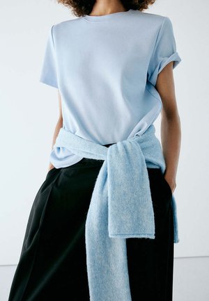 Personne portant une chemise bleu clair à manches courtes avec un pull assorti noué autour de la taille et un pantalon noir, les mains dans les poches.