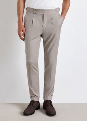 Man wearing light beige dress pants with pleats and dark brown lace-up shoes, standing with one hand in pocket against plain background.