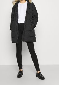 Abrigo acolchado negro con capucha forrada de piel, con cierre de cremallera y bolsillos laterales. Llevado sobre una camisa blanca con pantalones negros ajustados y mocasines.