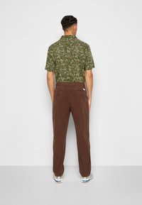 Man som står vänd bort med ryggen mot kameran, iklädd en grön kortärmad skjorta med lövmönster, bruna byxor och vita sneakers mot en enfärgad bakgrund.