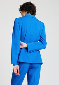 Person with short curly hair wearing a bright blue suit, standing with one arm bent behind their back against a plain background.