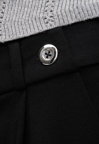 Bouton argenté à quatre trous fixé sur un tissu noir sous un bord de vêtement en maille grise texturée.
