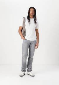 Weißes T-Shirt mit kurzen Ärmeln, graue Jeans und schwarz/weiße Sneaker. Hält eine hellgraue Umhängetasche und steht vor einem schlichten weißen Hintergrund.