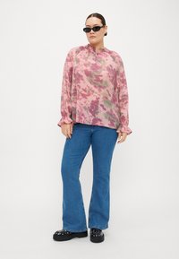 Blusa a maniche lunghe, a fantasia rosa e verde con collo arruffato, abbinata a jeans a zampa d'elefante blu e scarpe platform nere di grandi dimensioni.