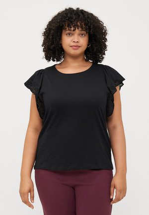 A woman with curly hair wears a black top featuring ruffled sleeves. She stands against a plain light background, looking at the camera.