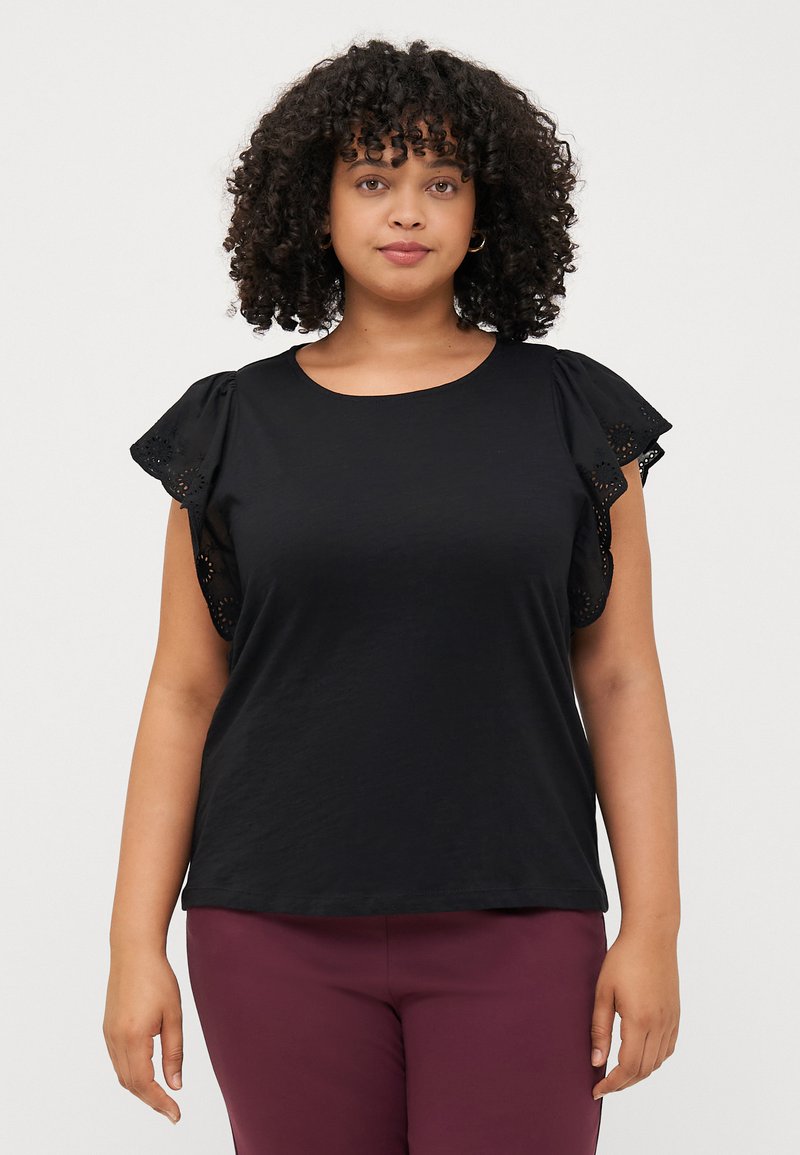 A woman with curly hair wears a black top featuring ruffled sleeves. She stands against a plain light background, looking at the camera.