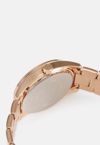Montre en métal rose gold avec un boîtier rond, une lunette texturée et un bracelet lisse. Comprend une couronne sur le côté et un dos polished en argent.