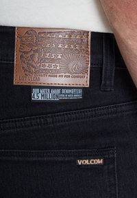 Zwarte denim jeans met een getextureerde bruine leren patch met een reliëfontwerp en logo's. Labels benadrukken waterbesparende productiemethoden.