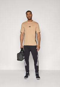 Beige sports T-shirt med sort logo, parret med sorte sweatpants med hvide striber. Modellen holder en sort træningstaske.