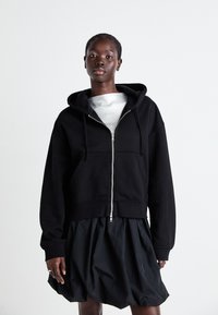 Felpa nera con zip e cappuccio, realizzata in tessuto morbido. Presenta un design cropped, ampie tasche anteriori e polsini a coste sopra una gonna scura.