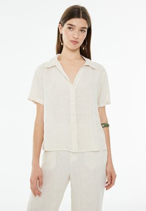 Camisa de lino beige claro de manga corta con cuello, botones en el frente y un ajuste relajado. Textura suave y diseño minimalista, adecuada para el uso casual.