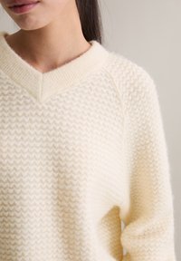 Sweter na drutach z dekoltem w serek w kolorze kremowym z teksturowanym wzorem w zygzak; posiada ściągacz na kołnierzu oraz miękki, puszysty materiał.