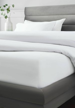 Kingsize-Bett mit weißer Bettwäsche und Kissen, grauem gepolstertem Rahmen und Kopfteil sowie einer Vase mit grünen Blättern im Hintergrund.