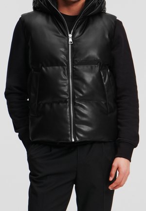 Homme portant une doudoune noire brillante avec une fermeture éclair frontale et une capuche, par-dessus une chemise noire à manches longues et un pantalon noir.