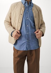 Beżowa pikowana kurtka na niebieskiej koszuli chambray, z zatrzaskami i luźnym krojem; brązowe spodnie dopełniają całość.