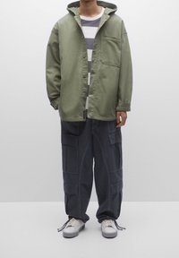 Chaqueta verde con capucha sobre una camiseta de rayas gris y blanca, combinada con pantalones cargo oscuros con múltiples bolsillos y zapatillas gris.