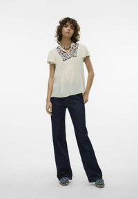 Hvid, kortærmet bluse med blomsterbroderi ved udskæringen; parret med mørkeblå flared jeans og blå, knyttede sandaler.