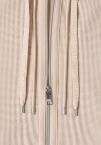 Nærbillede af beige ribstrikket hættetrøje med sølvlynlås og beige snore, der er prægede med teksten "OPDAG NYE STEDER".