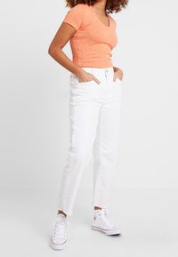 Vrouw in een oranje kortemouwen shirt, witte high-waist jeans met rafelige zomen, en witte high-top sneakers, met haar handen in de zakken.