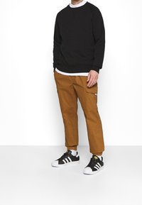 Sweatshirt noir à col rond, pantalon cargo marron avec poches latérales, et baskets noir et blanc avec un design à trois rayures.