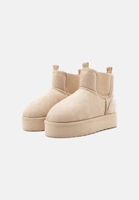 Bottines en daim beige avec un bout arrondi, une semelle plate, et des panneaux latéraux élastiques ; elles sont dotées de languettes en haut pour un enfilage facile.