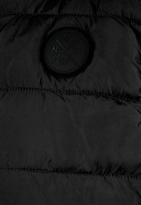 Fekete pufi kabát sima textúrával, varrott részletekkel és egy kör alakú logóval, amelynek közepén az "NMT 97" felirat található.