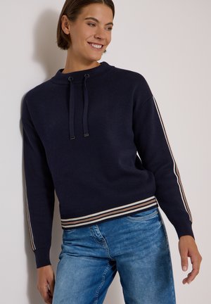 Femme aux cheveux courts et bruns, portant un pull bleu marine à poignets rayés et un jean bleu, souriante et appuyée contre un mur.