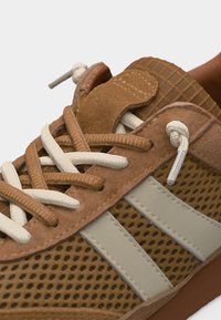 Κοντινό πλάνο καφέ αθλητικού παπουτσιού με ύφασμα από δίχτυ, δύο μπεζ ρίγες και κορδόνια ανάμεικτου μπεζ και καφέ χρώματος δεμένα σε κόμπο.