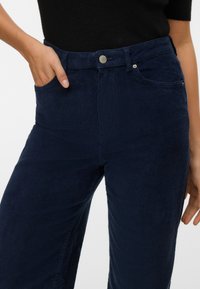 Femme portant un pantalon en velours côtelé bleu foncé avec la main dans la poche avant, associé à un haut noir.
