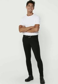 Black slim-fit pants with a belt and a white crew-neck t-shirt; model wearing black ankle boots; plain background.