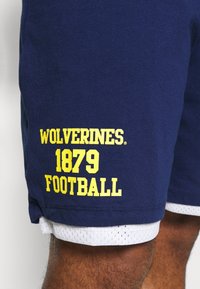 Mörkblå shorts med gul text "WOLVERINES 1879 FOOTBALL" som bärs över vita nätshorts på en persons ben.