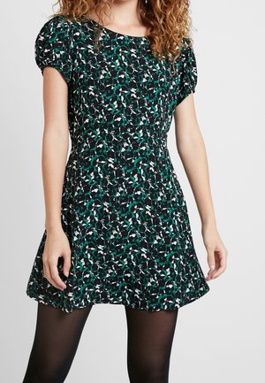 Vrouw draagt een korte zwarte jurk met groen-wit bloemenpatroon en pofmouwtjes, gecombineerd met zwarte doorschijnende panty's.