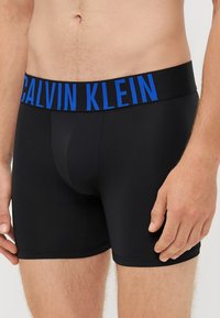 Boxeri scurți din bumbac negru cu un logo al Calvin Klein de culoare albastră pe talie, având un design ajustat și o textură netedă.