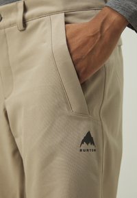 Pantalons beiges avec une texture lisse, dotés d'une poche latérale et d'un logo de montagne noir portant l'inscription "BURTON" sur la cuisse.