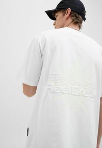 Hvid bomulds T-shirt med et tekstureret logo "ReelBreak" og et sol-design i en lidt lysere nuance, korte ærmer, afslappet pasform.