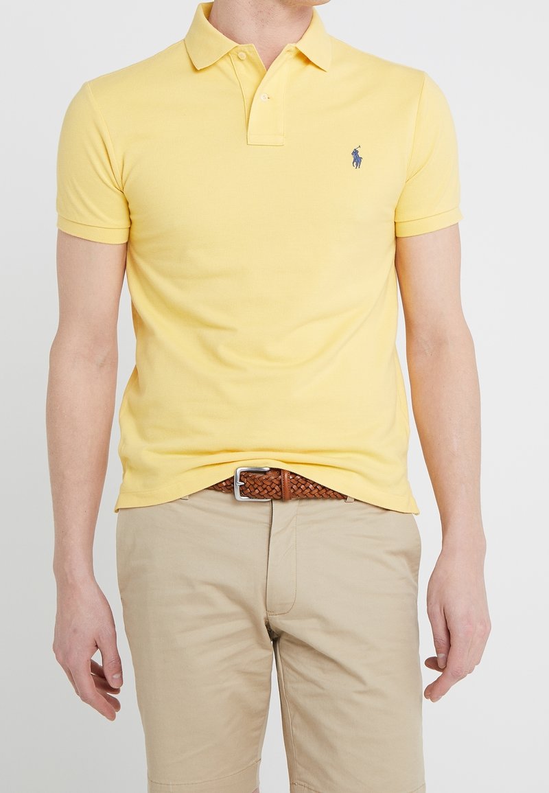 Homme portant un polo jaune à manches courtes avec un logo bleu, un short beige et une ceinture tressée marron, debout devant un fond uni.
