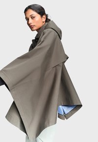 Femme aux cheveux foncés tirés en arrière, portant une cape ample taupe à capuche avec une doublure bleue, debout devant un fond clair uni.