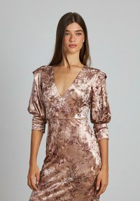Donna con lunghi capelli castani, indossa un lucido vestito in velluto floreale marrone con scollatura a V profonda e maniche a sbuffo di tre quarti, in piedi contro uno sfondo semplice.