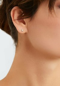 Pequeño pendiente de botón redondo en metal plateado. Diseño sencillo con una superficie reflectante. Lleva en el lóbulo de la oreja contra un tono de piel neutro.