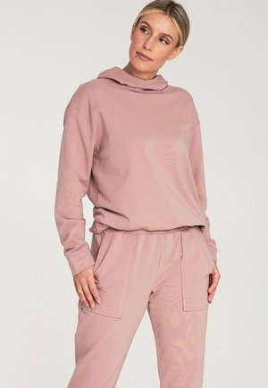 Jasnoróżowy, długi sweter z rękawami, z wysokim, złożonym kołnierzem oraz luźnym krojem. Wykonany z miękkiego materiału, z opuszczonymi ramionami. Widać dołączone spodnie.
