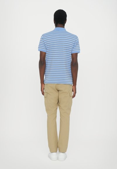 Ριγέ μπλε πόλο μπλούζα με κοντό μανίκι, συνδυασμένη με μπεζ cargo παντελόνι. Μοντέλο στραμμένο πλάτη, φορώντας άσπρα αθλητικά παπούτσια.