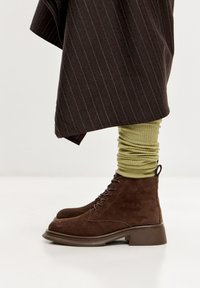 Botines marrones de ante con cordones, usados con calcetines de canalé verdes y una falda o abrigo de rayas marrones visible desde la rodilla hacia abajo.