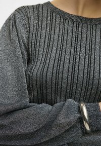 Pullover nero metallizzato con pieghe verticali, tessuto testurizzato e scollatura rotonda. Maniche lisce che si raccolgono al polsino con un bracciale argentato lucido.