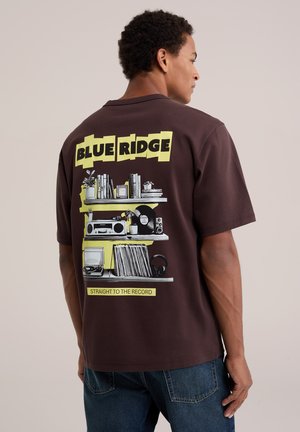Homme portant un t-shirt marron foncé avec texte jaune "BLUE RIDGE" et graphique d'étagères contenant des livres, des plantes, une chaîne stéréo, des disques vinyles et des écouteurs.