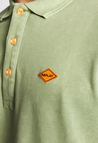 Jasnozielona koszulka polo z pomarańczowymi guzikami i diamentowym pomarańczowym naszywką z logo "REPLAY". Teksturowana tkanina.