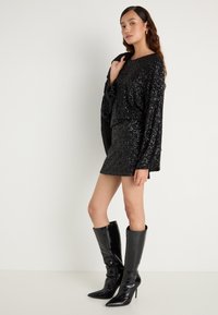 Juoda blizganti suknelė su ilgomis rankovėmis ir aptemptu dizainu, derinama su juodais odiniais stiletto batais iki kelių. Paprastas fonas.