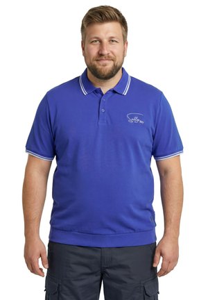 Uomo barbuto in piedi, che indossa una maglietta polo blu con il logo "Salty Boy" e pantaloncini cargo scuri, su uno sfondo bianco uniforme.