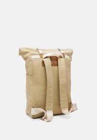 Beige roll-top rugzak gemaakt van stevig stof. Heeft witte banden, leren accenten, twee zijvakken en een verstevigde bodem.