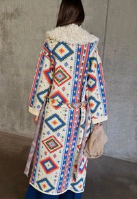 Γυναίκα που φοράει μακρύ, πολύχρωμο παλτό με γεωμετρικό σχέδιο και άσπρο γιακά από φίδι, κρατώντας μπεζ σουέντ τσάντα, στέκεται μπροστά σε τοίχο από σκυρόδεμα.